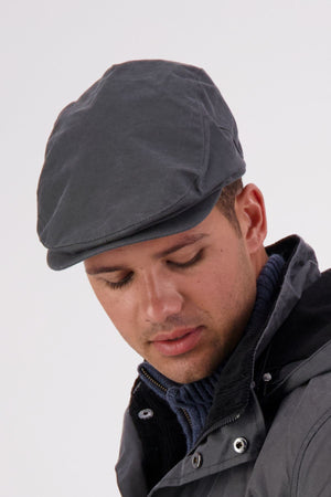 A man wearing the Swanndri Birmingham Oilskin Flat Cap and a gray waxed jacket looks down with a neutral expression against a light background.