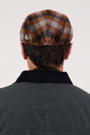 A person is shown from behind wearing a Swanndri Bristol Flat Cap in Teddington Check and a dark jacket with a black collar against a plain, light-colored background.