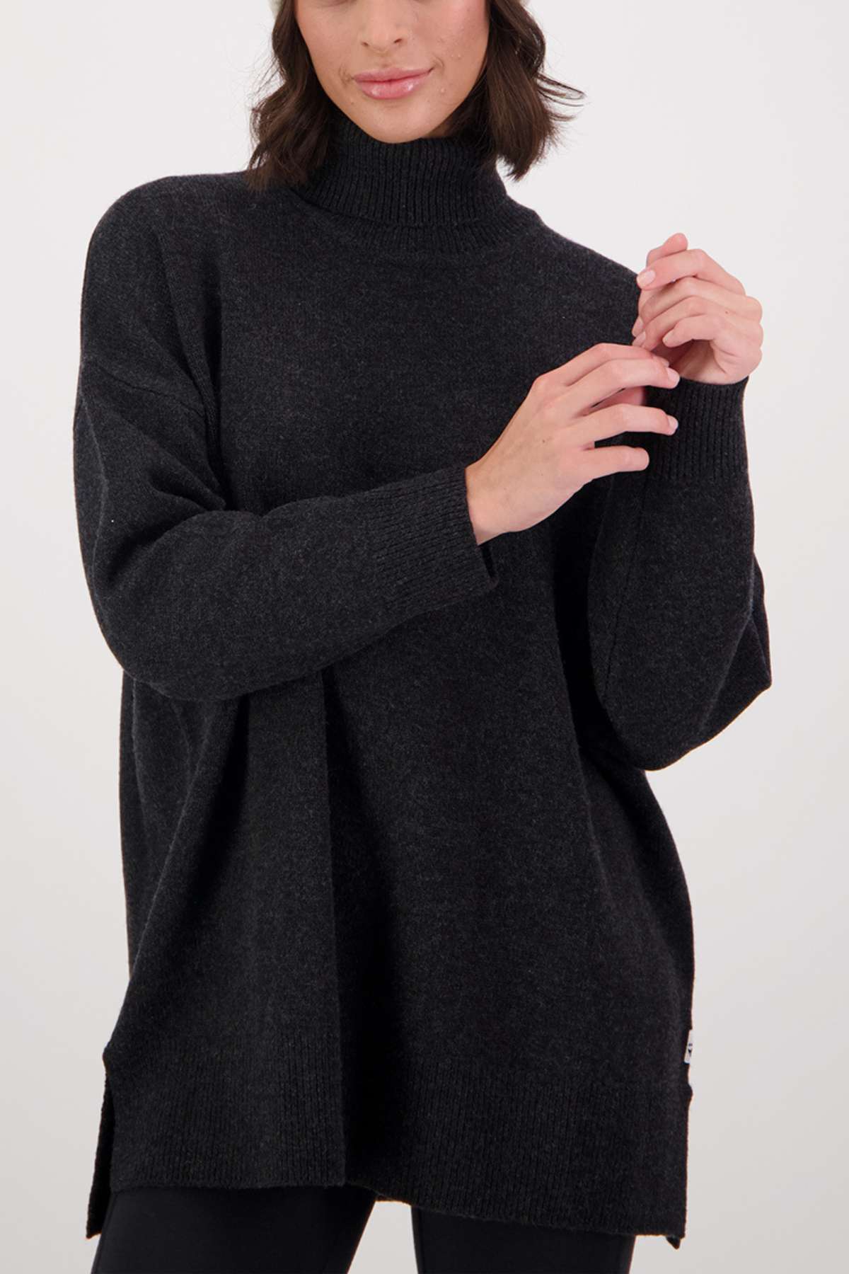 A woman wearing the Swanndri Brunner Merino Roll Neck Knit in a loose, dark fit and black leggings stands against a plain white background, her hands gently clasped near her chest.