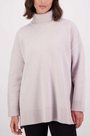 A woman wearing the Swanndri Brunner Merino Roll Neck Knit in light lavender, featuring long sleeves with ribbed cuffs and hem, stands against a plain white background.