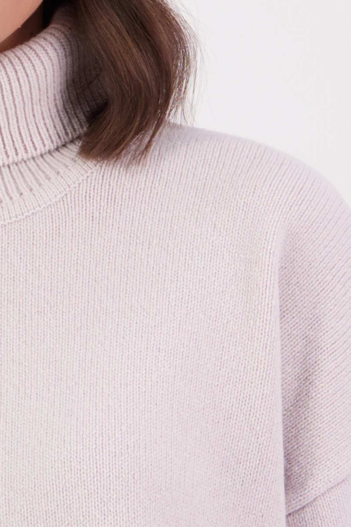 Close-up of a person wearing the Swanndri Brunner Merino Roll Neck Knit in a light color. Only the lower face, neck, shoulder, and some brown hair are visible against a plain, light background.