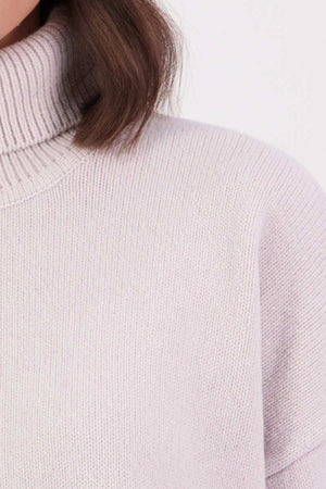 Close-up of a person wearing the Swanndri Brunner Merino Roll Neck Knit in a light color. Only the lower face, neck, shoulder, and some brown hair are visible against a plain, light background.