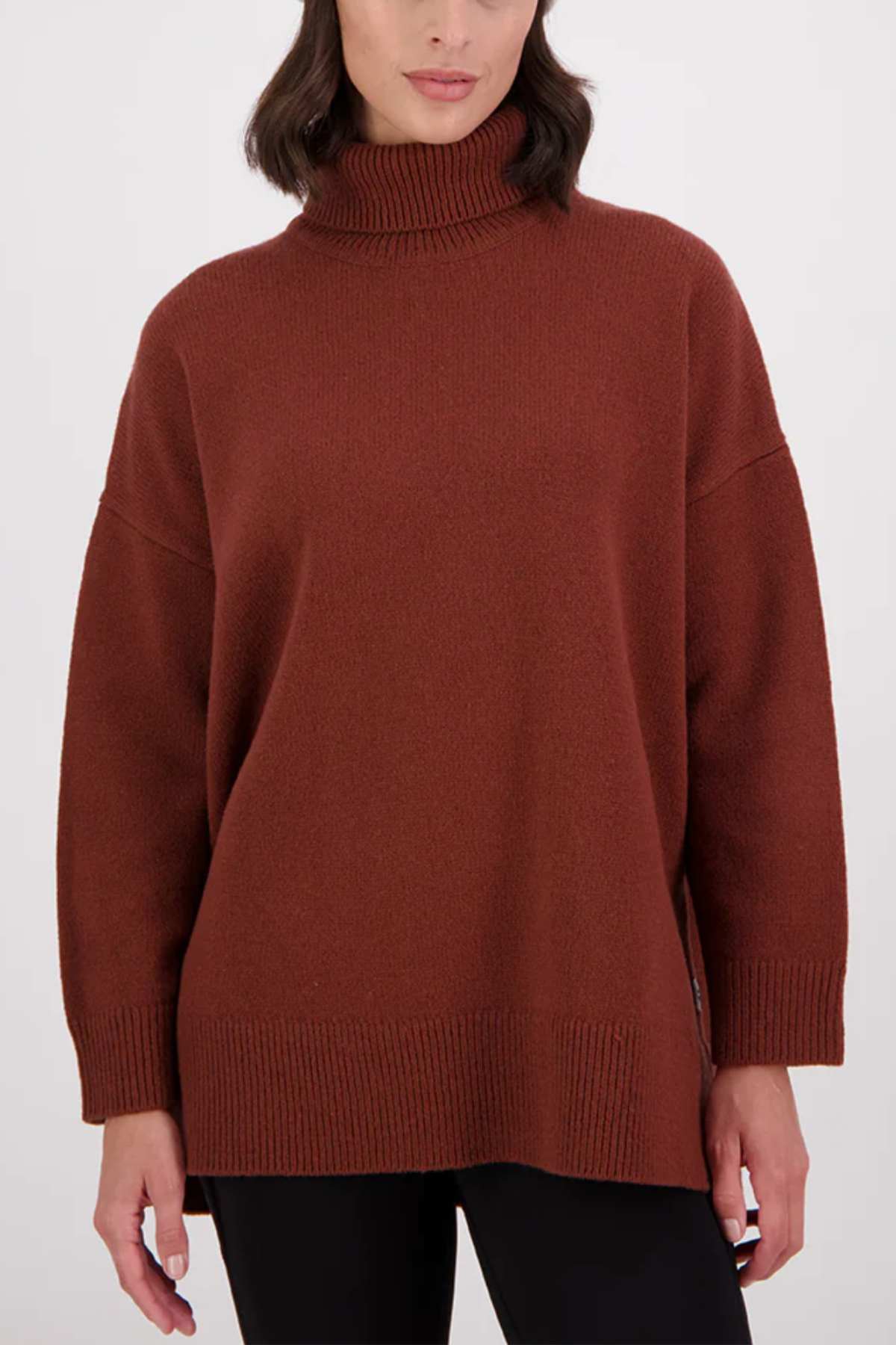 A woman wears the Swanndri Brunner Merino Roll Neck Knit, a loose-fitting brown sweater with ribbed cuffs, hem, and neckline, styled with black pants against a plain light background.