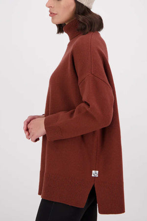 A woman in profile wears a Swanndri Brunner Merino Roll Neck Knit sweater, a beige knit hat, and black pants. She has shoulder-length dark hair and stands against a plain background.
