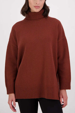 A woman wears the Swanndri Brunner Merino Roll Neck Knit, a loose-fitting brown sweater with ribbed cuffs, hem, and neckline, styled with black pants against a plain light background.