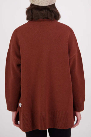 A person wearing a beige knit beanie and an oversized, long-sleeve rust Swanndri Brunner Merino Roll Neck Knit sweater, shown from the back against a plain light background.