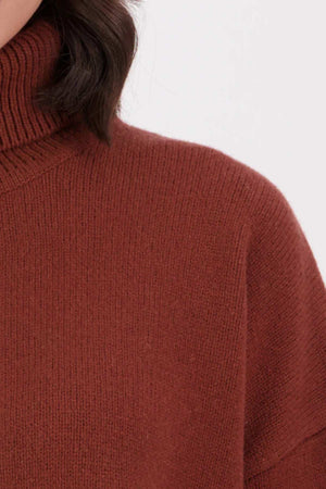 Close-up of a person wearing the Swanndri Brunner Merino Roll Neck Knit in rust. The image shows part of the shoulder, neck, and some dark wavy hair against a plain light background.
