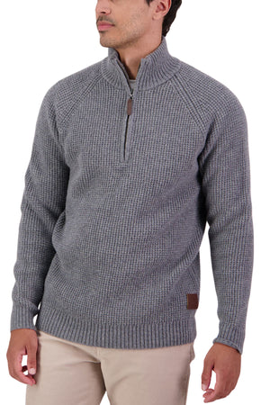 A man stands looking to the side, wearing a Swanndri Chalky Island Waffle Jumper in gray with long sleeves and a half-zip collar, paired with tan pants.