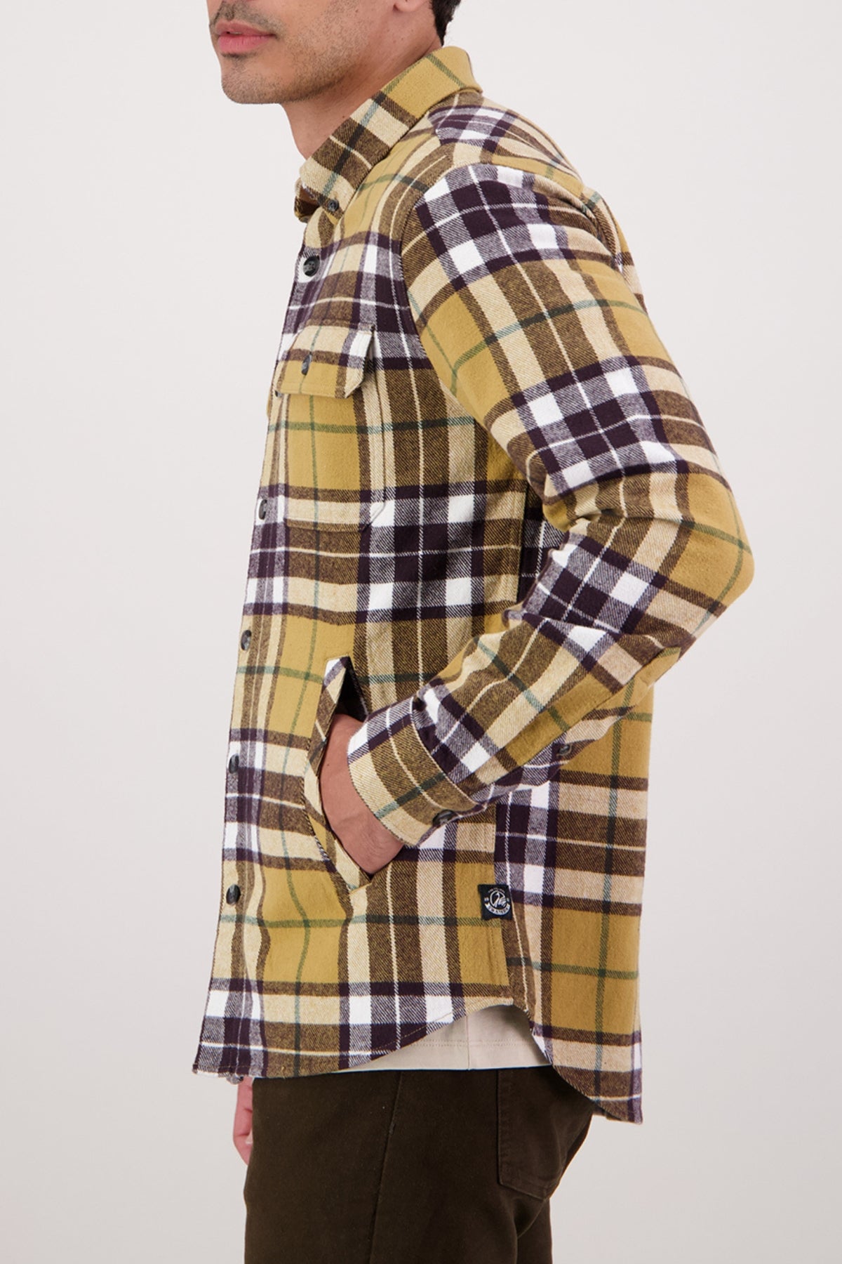 Wearing the Swanndri Crofton Shirt in a yellow, brown, and white plaid inspired by heritage workwear, made from 100% cotton with chest and side pockets, they stand sideways with a hand in their pocket against a plain light background.