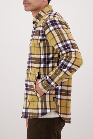 Wearing the Swanndri Crofton Shirt in a yellow, brown, and white plaid inspired by heritage workwear, made from 100% cotton with chest and side pockets, they stand sideways with a hand in their pocket against a plain light background.