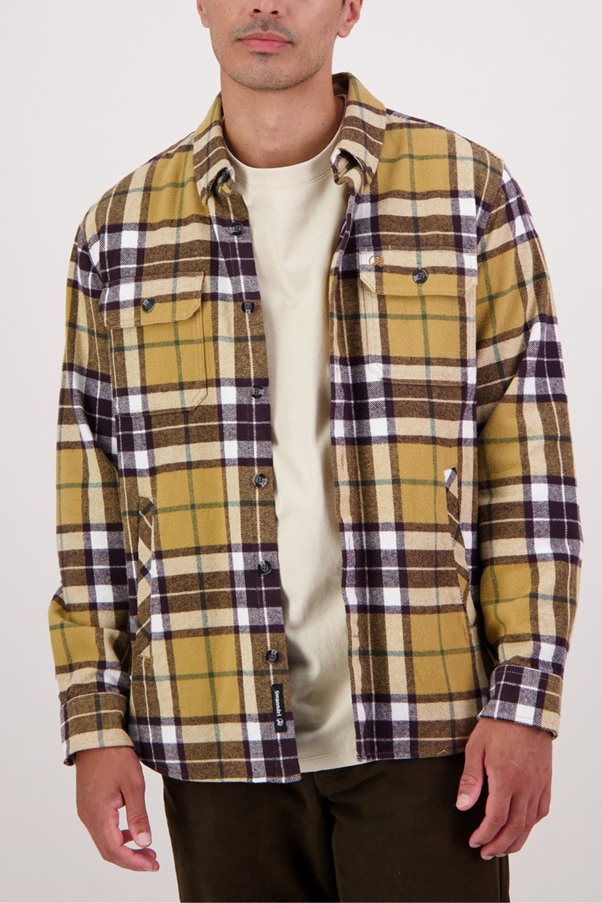 Wearing a Swanndri Crofton Shirt—100% cotton yellow, brown, and white plaid—over a beige tee with dark brown pants, this person stands against a light backdrop for a classic heritage workwear style.