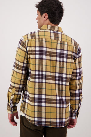 A man is shown from behind wearing the Swanndri Crofton Shirt, featuring yellow and brown plaid in 100% cotton, paired with dark pants and standing against a plain, light background.