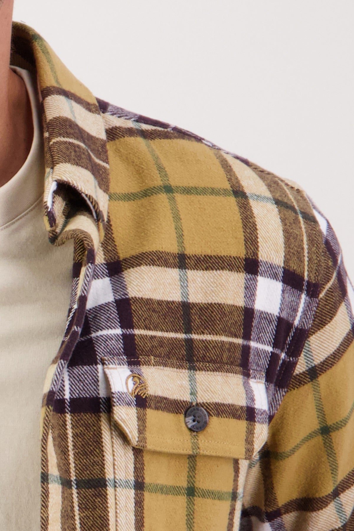 A close-up of a person wearing the Swanndri Crofton Shirt—a plaid flannel in yellow, brown, white, and green 100% cotton—over a light t-shirt. The shirt features a buttoned front pocket; only the upper torso is shown.