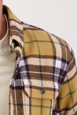 A close-up of a person wearing the Swanndri Crofton Shirt—a plaid flannel in yellow, brown, white, and green 100% cotton—over a light t-shirt. The shirt features a buttoned front pocket; only the upper torso is shown.