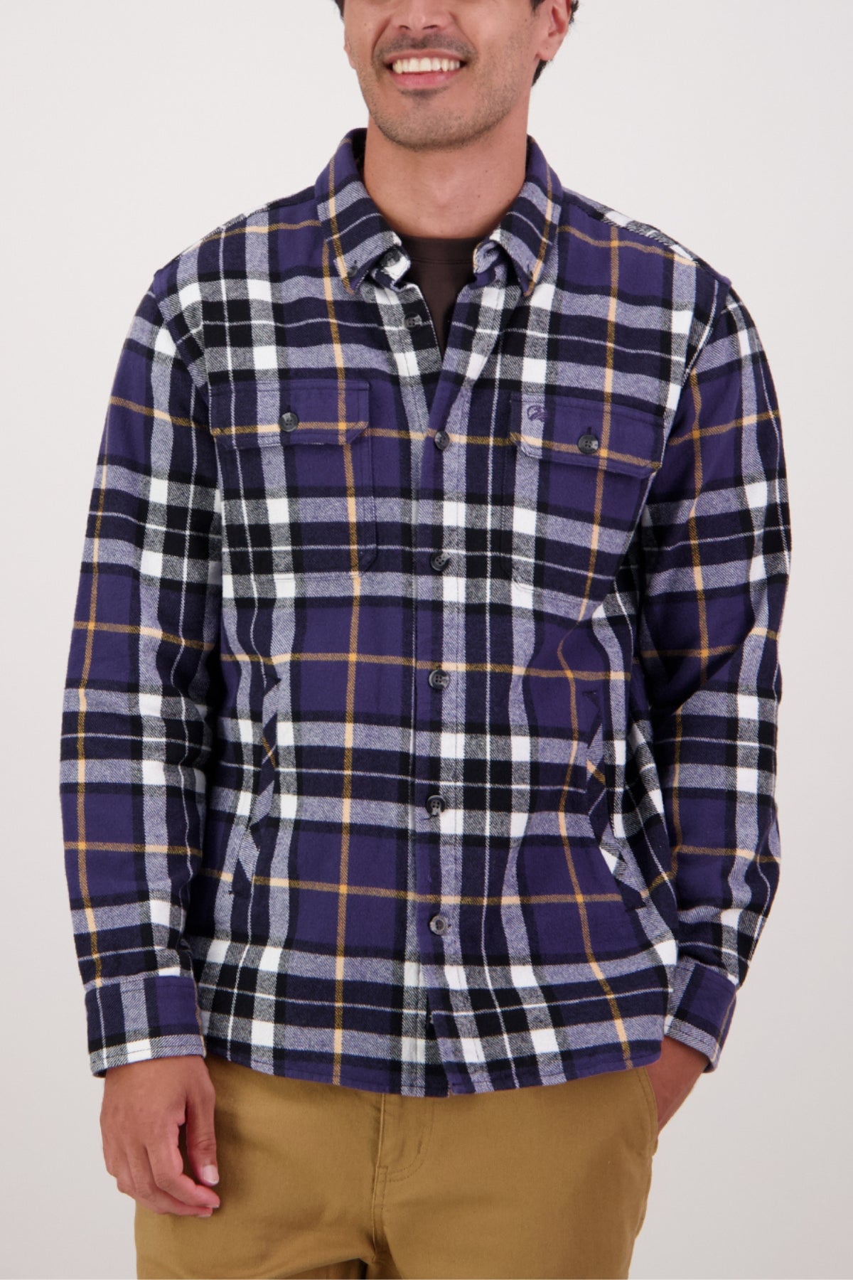 A person in a Swanndri Crofton Shirt featuring purple, black, white, and yellow plaid with tan pants, smiling with one hand by their side against a plain light background. The shirt is made from 100% cotton.