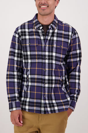 A person in a Swanndri Crofton Shirt featuring purple, black, white, and yellow plaid with tan pants, smiling with one hand by their side against a plain light background. The shirt is made from 100% cotton.