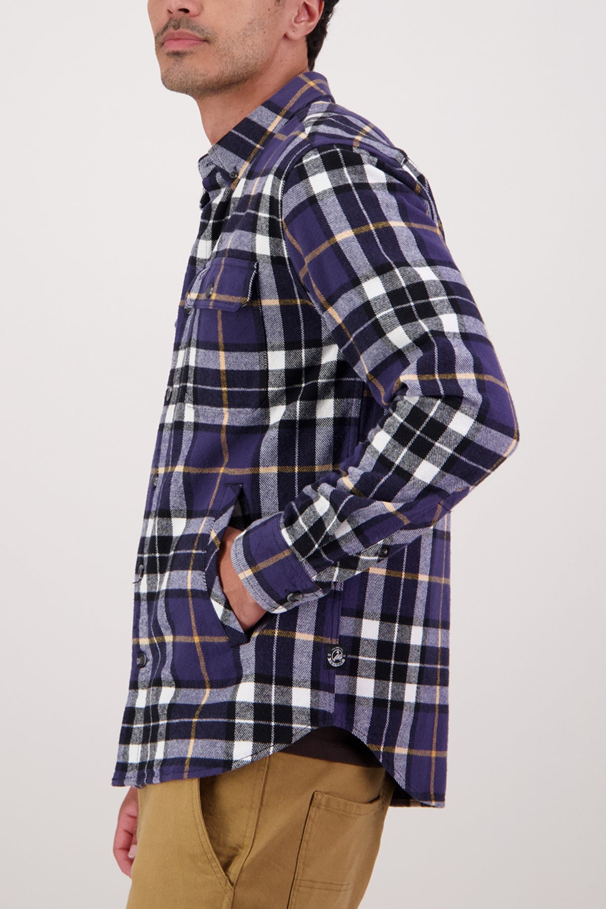 A person stands sideways with a hand in their tan pants pocket, wearing the Swanndri Crofton Shirt—a plaid 100% cotton button-up inspired by heritage workwear. The background is plain and light-colored.