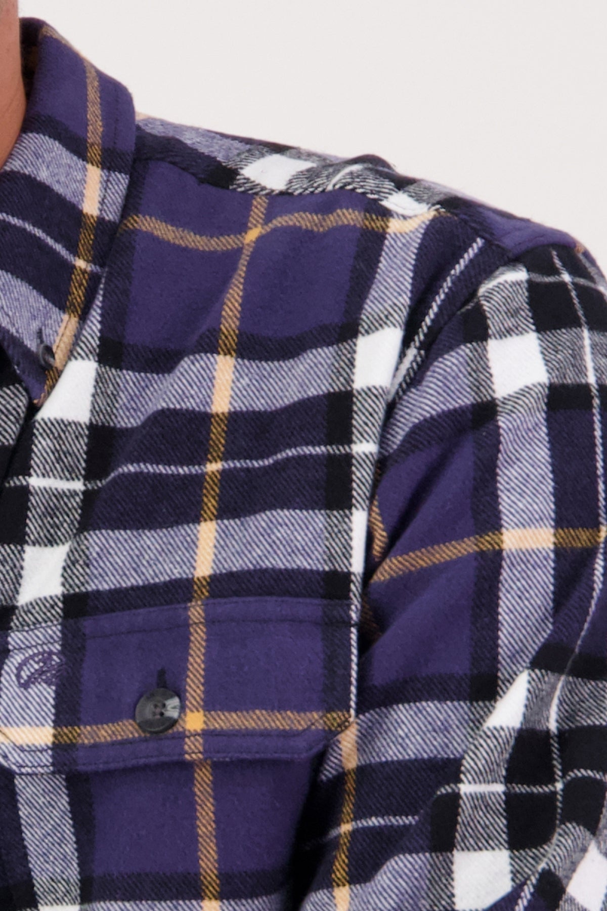 A close-up shows the upper chest and shoulder of someone wearing the Swanndri Crofton Shirt, a heritage-inspired flannel in purple, black, white, and yellow plaid with a buttoned front pocket. Made from 100% cotton.