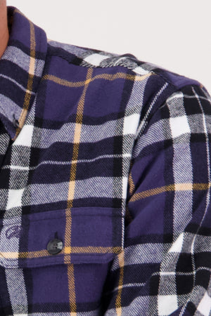 A close-up shows the upper chest and shoulder of someone wearing the Swanndri Crofton Shirt, a heritage-inspired flannel in purple, black, white, and yellow plaid with a buttoned front pocket. Made from 100% cotton.