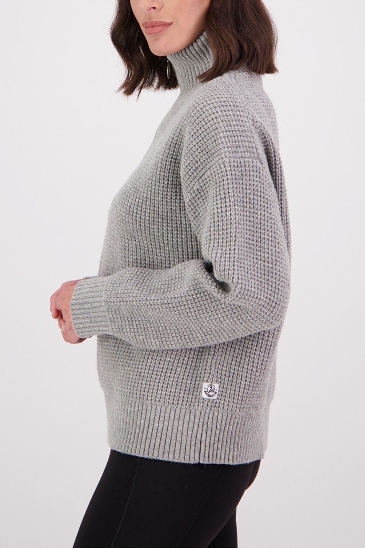 A woman with shoulder-length brown hair models the Swanndri Darlinghurst Zip Jumper in light gray, featuring a ribbed lambswool design and oversized collar, paired with black pants against a white background.