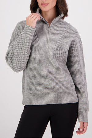 A woman wears a Swanndri Darlinghurst Zip Jumper—a light gray lambswool pullover with an oversized collar and long sleeves—paired with black pants. Only the lower part of her face is visible.