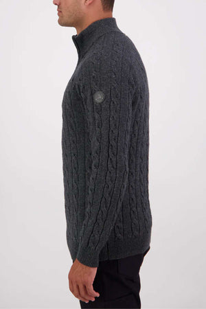 A man in profile wears a dark gray Swanndri Doncaster v2 Zip Cable Knit sweater with a quarter-zip collar and a small circular patch on the upper sleeve, standing against a plain light background.