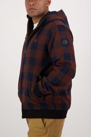 A person wearing a Swanndri Dunwich Wool Hoody in brown and navy checkered merino wool with a hood, standing sideways with hands in their pockets. They have on tan-colored pants. The background is plain white.