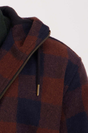 Close-up of a person's upper body wearing the Swanndri Dunwich Wool Hoody, featuring a large red and navy checkered pattern. This insulated hoody has a partially visible zipper and a black hood string hanging down. The background is a neutral, light color.