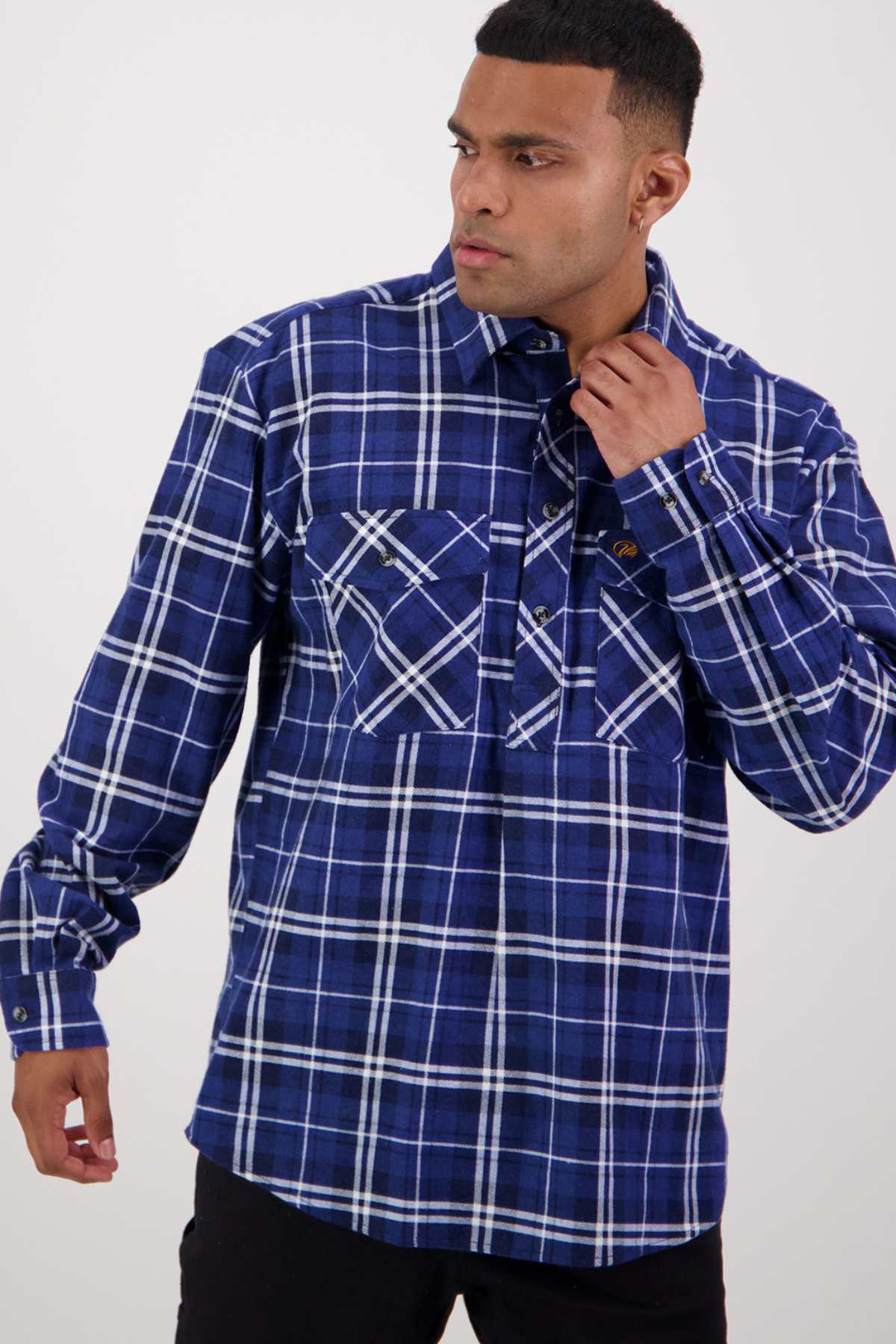 A person wearing a Swanndri Egmont Flannelette Twin Pack shirt in a blue and white plaid pattern looks to the side while adjusting their collar. They have short, dark hair and a neutral expression, with their left hand touching the shirt and the right hand by their side. The background is plain and light-colored.