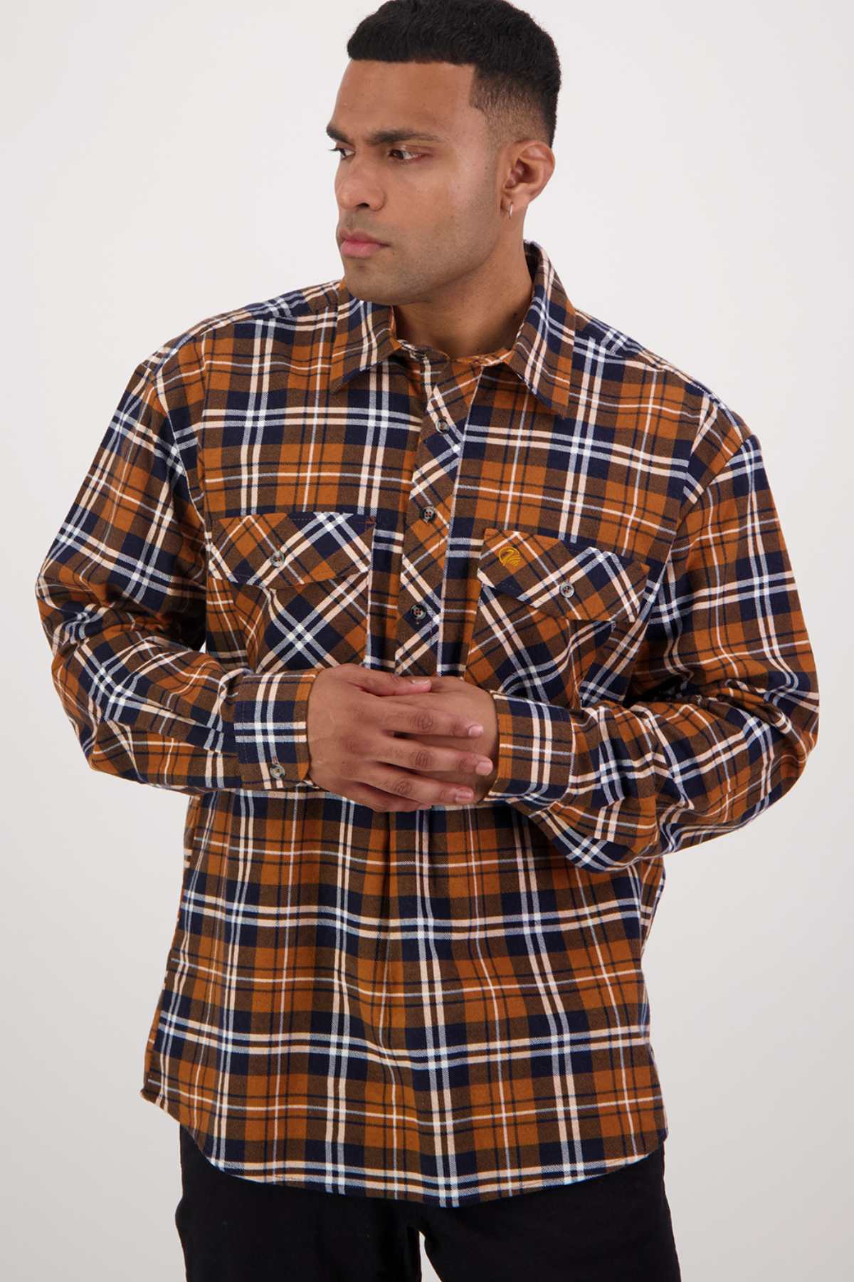A man stands against a plain background, wearing a long-sleeved, button-up plaid shirt in brown, navy blue, and white brushed cotton from the Swanndri Egmont Flannelette Twin Pack. He has his hands clasped in front of him and is looking to his right with a serious expression.