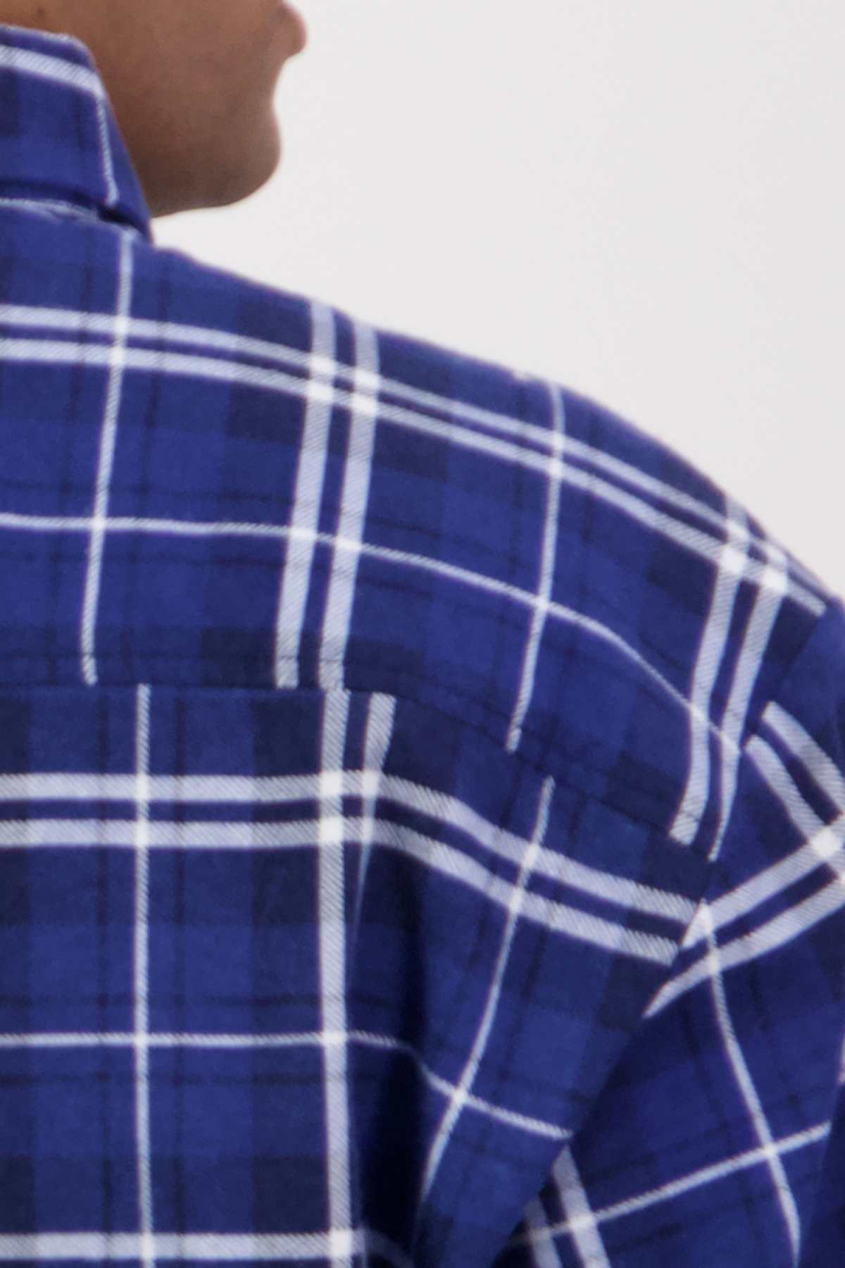 A close-up image showing the shoulder and part of the back of a person wearing a shirt from the Swanndri Egmont Flannelette Twin Pack. The fabric pattern includes intersecting white lines creating a grid-like design on a blue background. Made from soft, brushed cotton flannelette, the person's face is not visible.