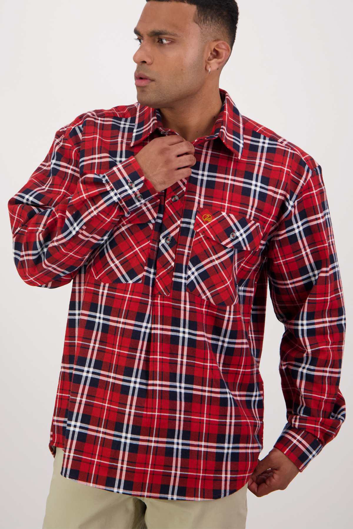 A man wearing a red, white, and black plaid shirt from the Swanndri Egmont Flannelette Twin Pack poses against a neutral background. He holds the collar of his long sleeve shirt with one hand and looks off to the side. He has short hair and is dressed in light-colored pants.