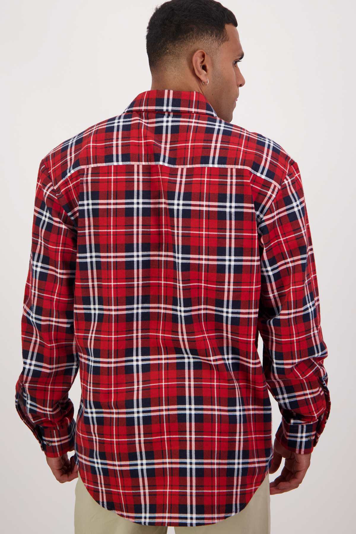 A person stands with their back to the camera, showcasing a Swanndri Egmont Flannelette Twin Pack long-sleeve shirt in a red, white, and black plaid pattern. The shirt features a classic design with intersecting lines, and the individual complements it with light-colored pants against a plain white background.