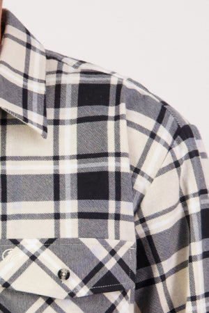 A close-up of the Swanndri Egmont Flannelette Twin Pack, featuring a black, white, and beige plaid shirt in soft brushed cotton. The image highlights the collar, shoulder, and buttoned chest pocket with intersecting lines and squares.