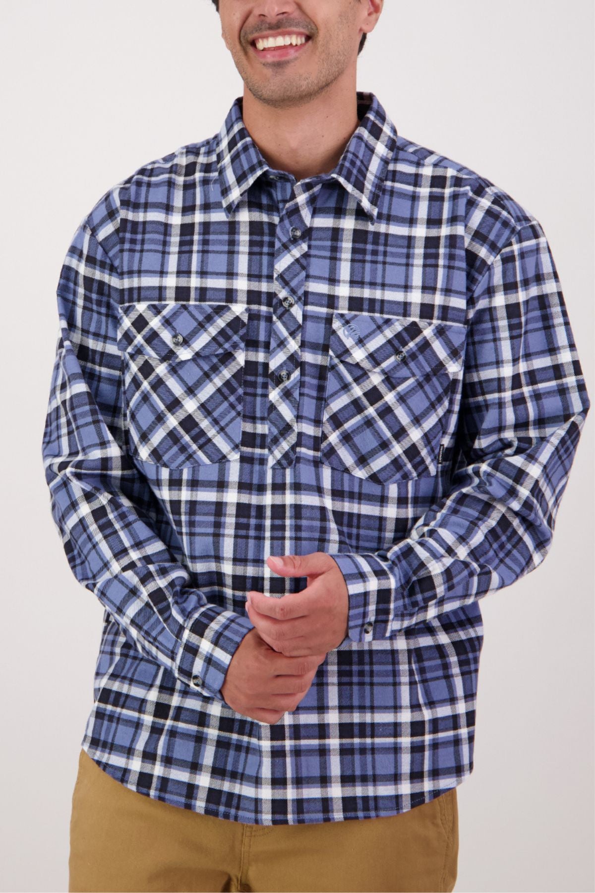 A person smiles while wearing a Swanndri Egmont Flannelette Twin Pack blue and black plaid button-up shirt with chest pockets and tan pants, posing against a plain light background.