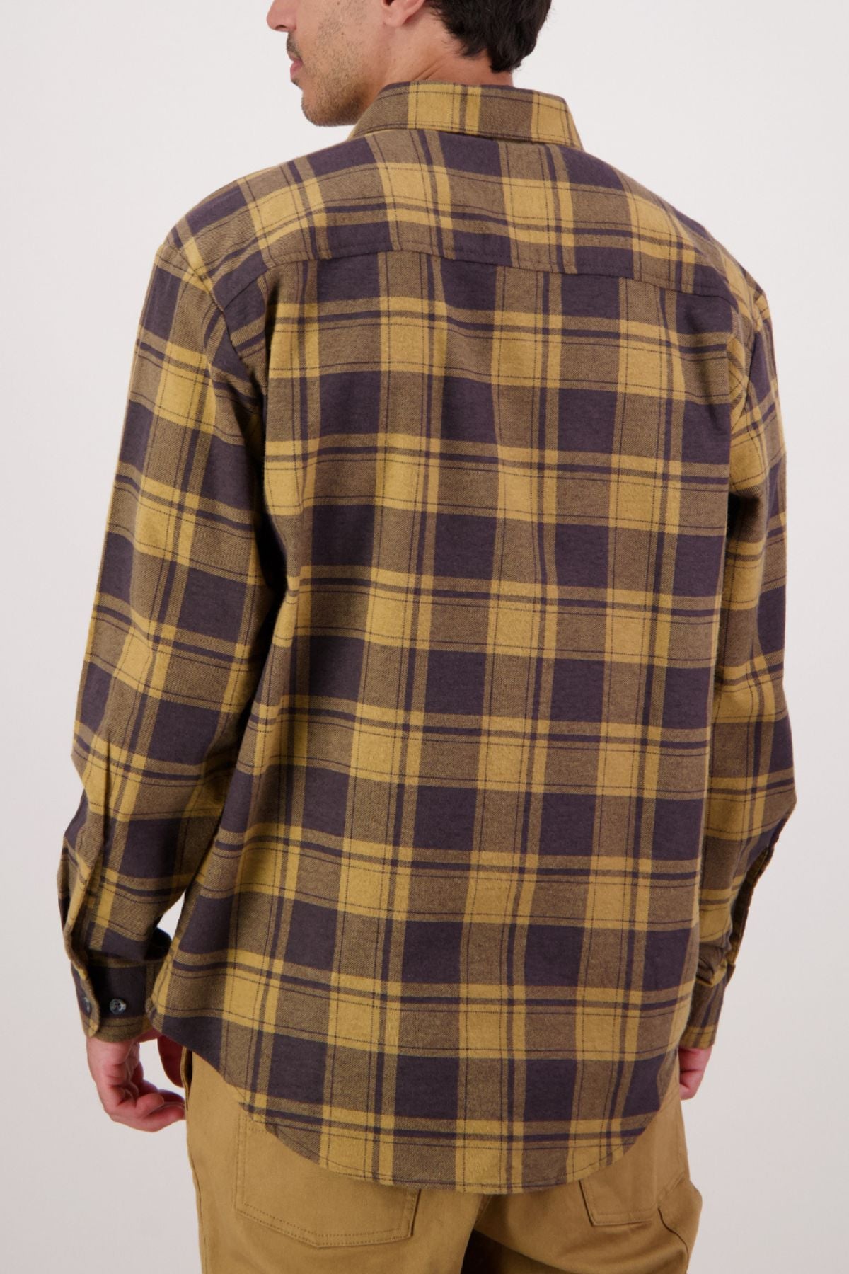 A person is seen from behind wearing a yellow and brown plaid Swanndri Egmont Flannelette Twin Pack long sleeve shirt with tan pants, standing against a plain light background.