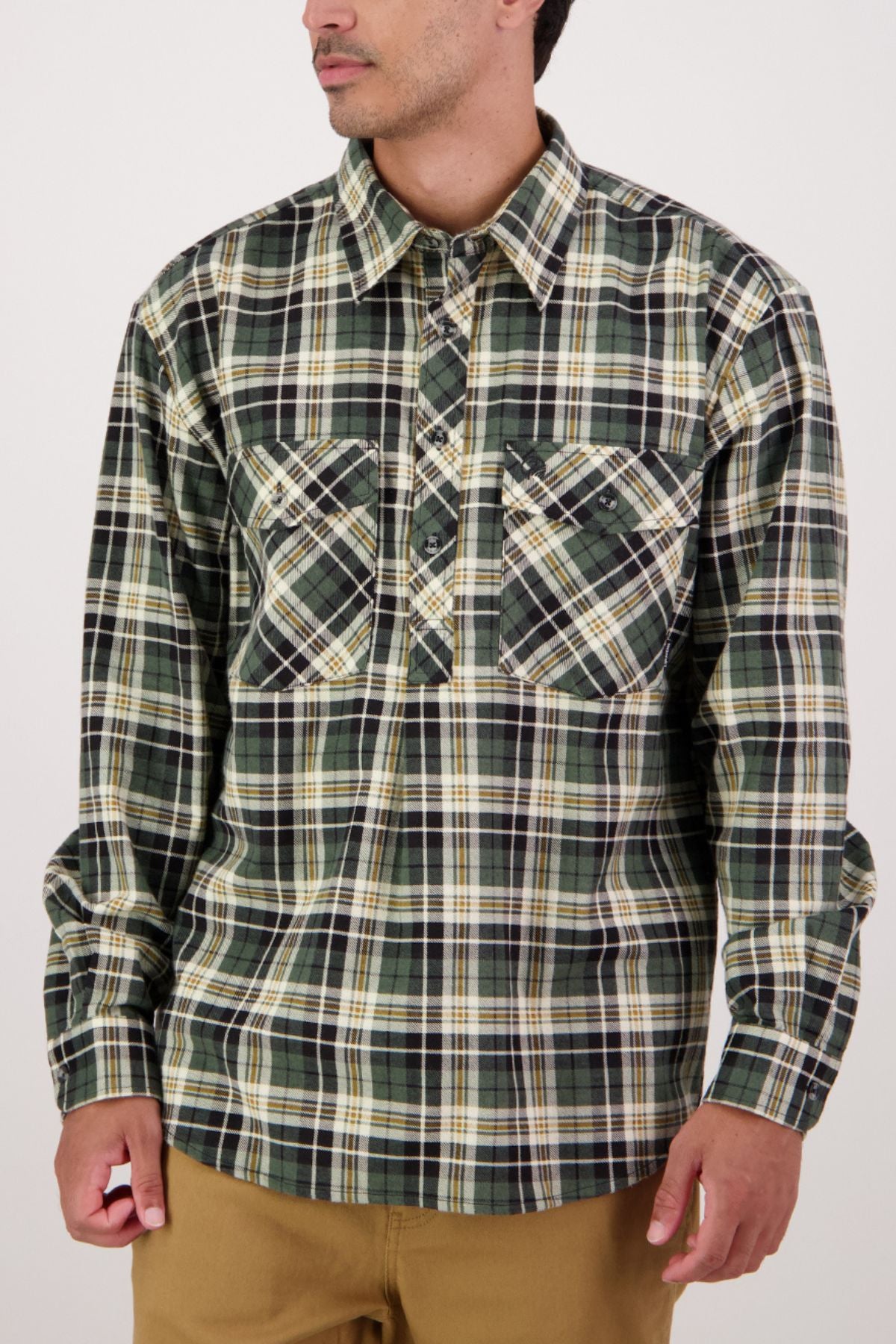 A man, arms relaxed and looking left, wears a Swanndri Egmont Flannelette Twin Pack shirt—a heavy long-sleeve in green, black, white, and yellow plaid with two chest pockets.