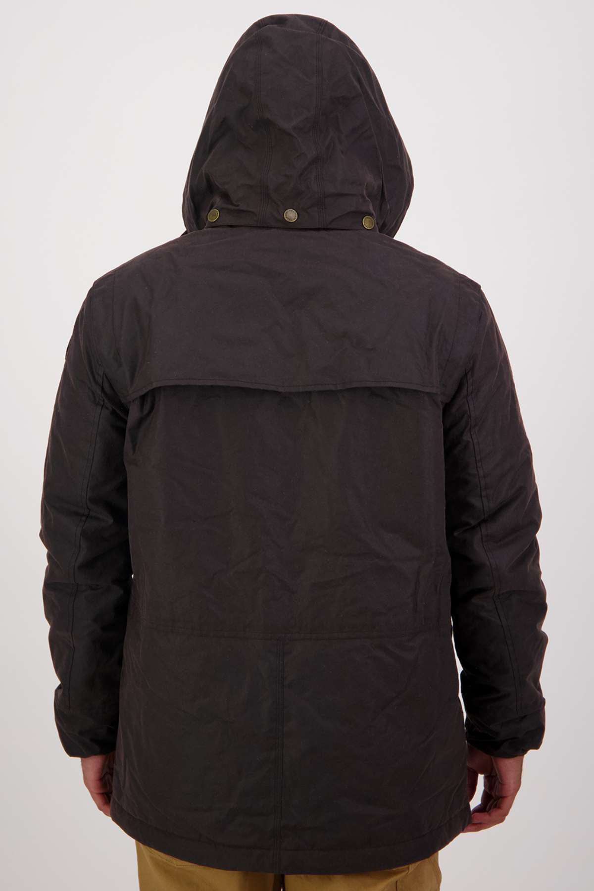 A person is seen from behind wearing a Swanndri Evans Flat Oilskin Jacket with the hood up and brown pants. The jacket has a flap across the upper back and visible seams, standing out against a simple, light background.