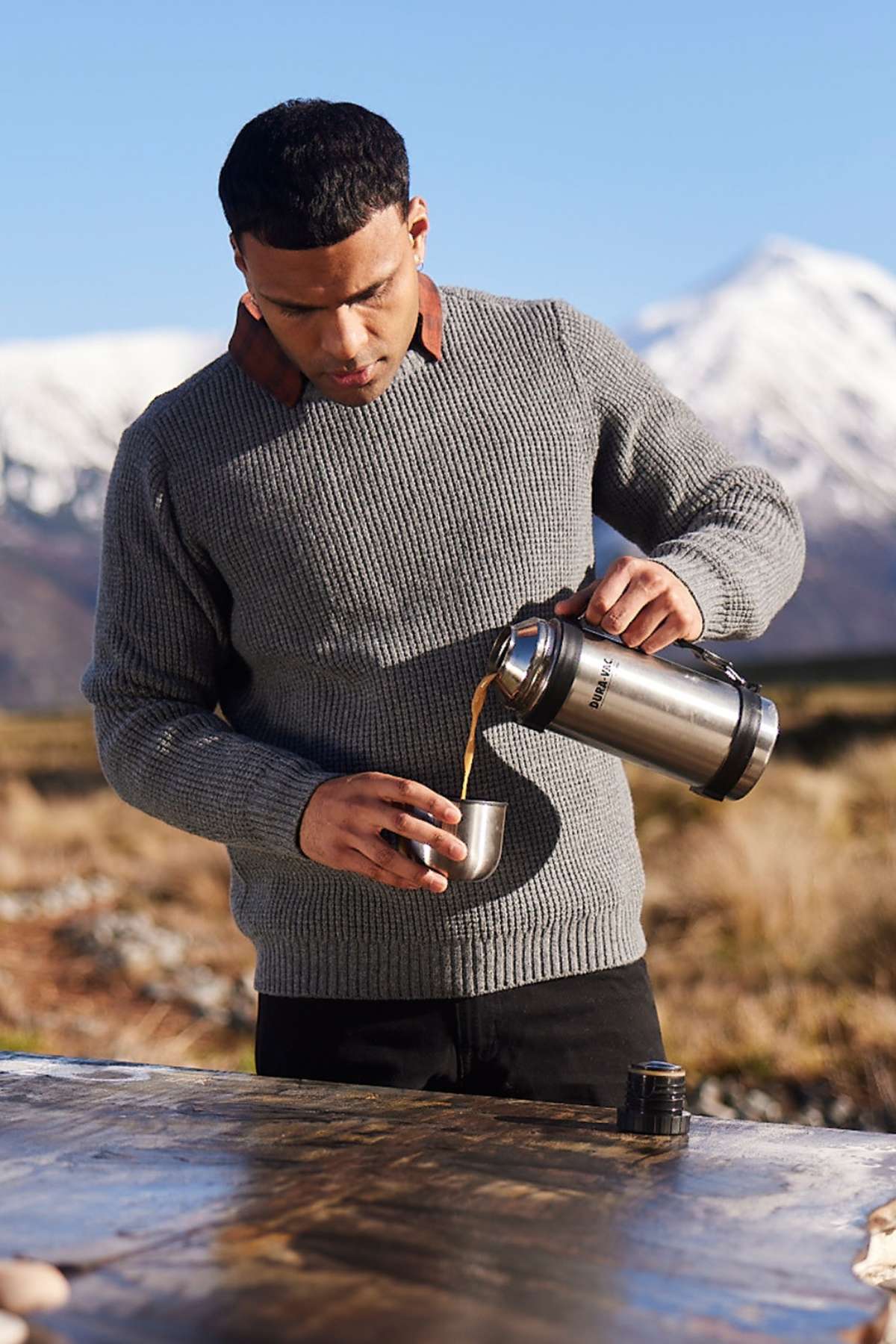 A man in a Swanndri Fistral Waffle Knit gray sweater pours a hot beverage from a thermos into a cup outdoors, against the backdrop of snow-capped mountains and clear blue skies, standing by a wooden surface in this rugged natural landscape.