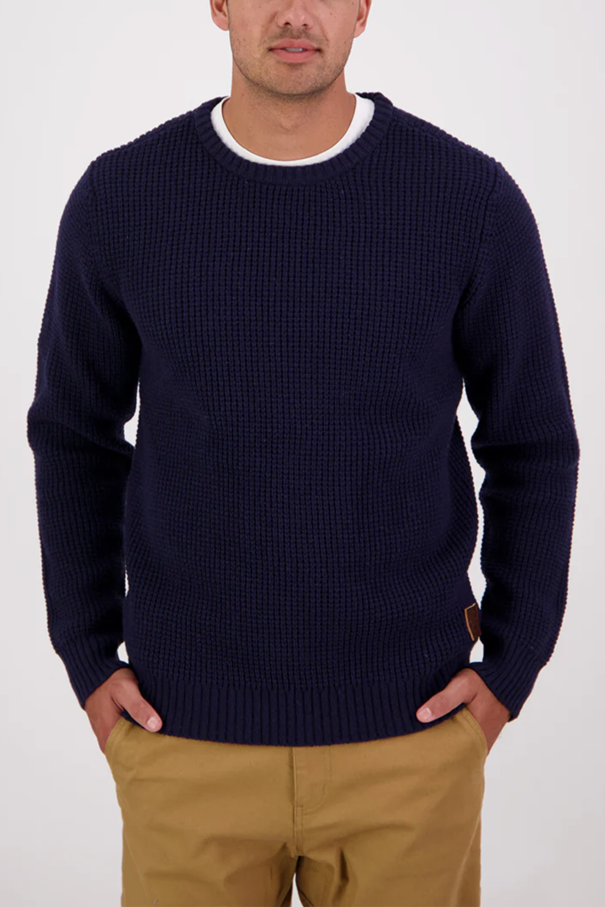 A man wears the Swanndri Fistral Waffle Knit navy sweater, with a subtle waffle pattern, layered over a white t-shirt and tan pants, standing with hands in pockets against a plain white background.