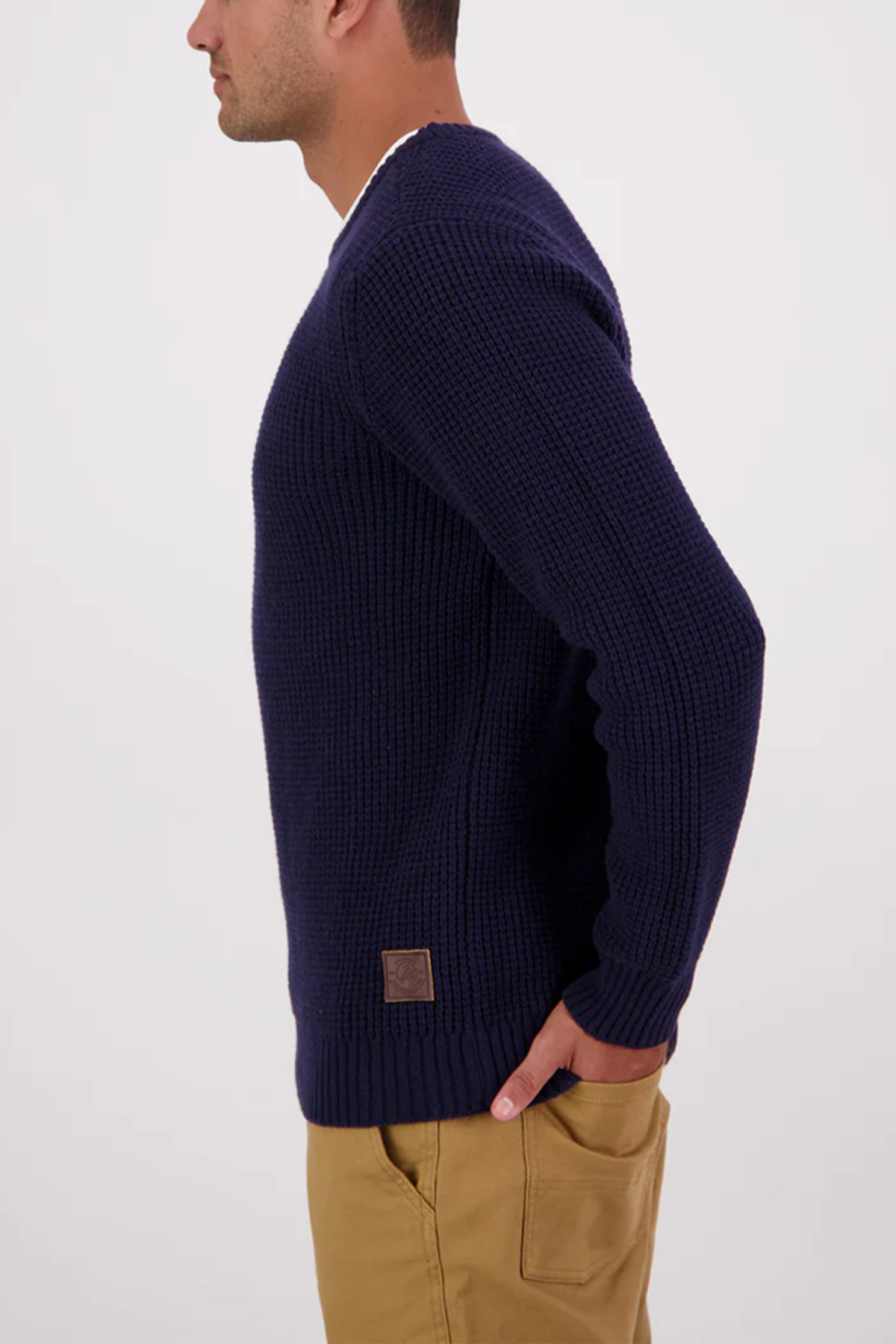 A man in profile wears the Swanndri Fistral Waffle Knit in navy blue with tan pants, one hand in his pocket, highlighting its soft lambswool and textured waffle knit design against a plain white background.