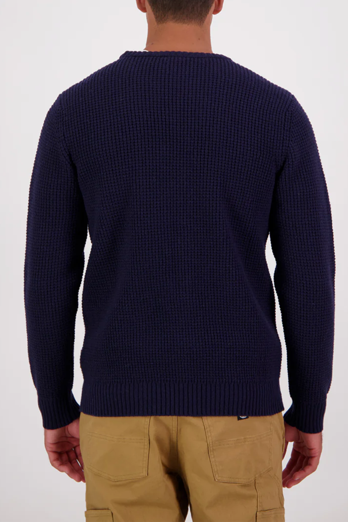 A person faces away from the camera, wearing the Swanndri Fistral Waffle Knit sweater in dark navy and tan pants, set against a plain white background.