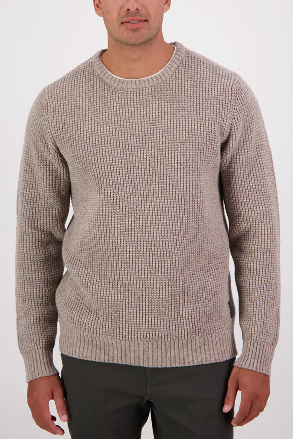 A man stands against a plain white background wearing the Swanndri Fistral Waffle Knit, a light beige lambswool crew-neck sweater with a subtle waffle design, paired with dark pants and his arms relaxed by his sides.