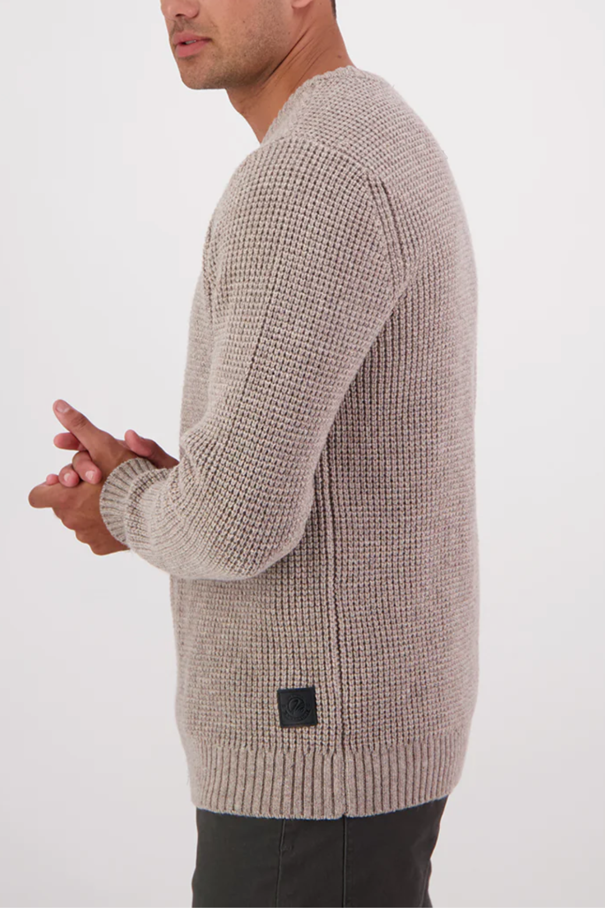 A man in a light beige Swanndri Fistral Waffle Knit sweater with a subtle textured design and dark pants stands sideways against a white background, hands clasped in front.
