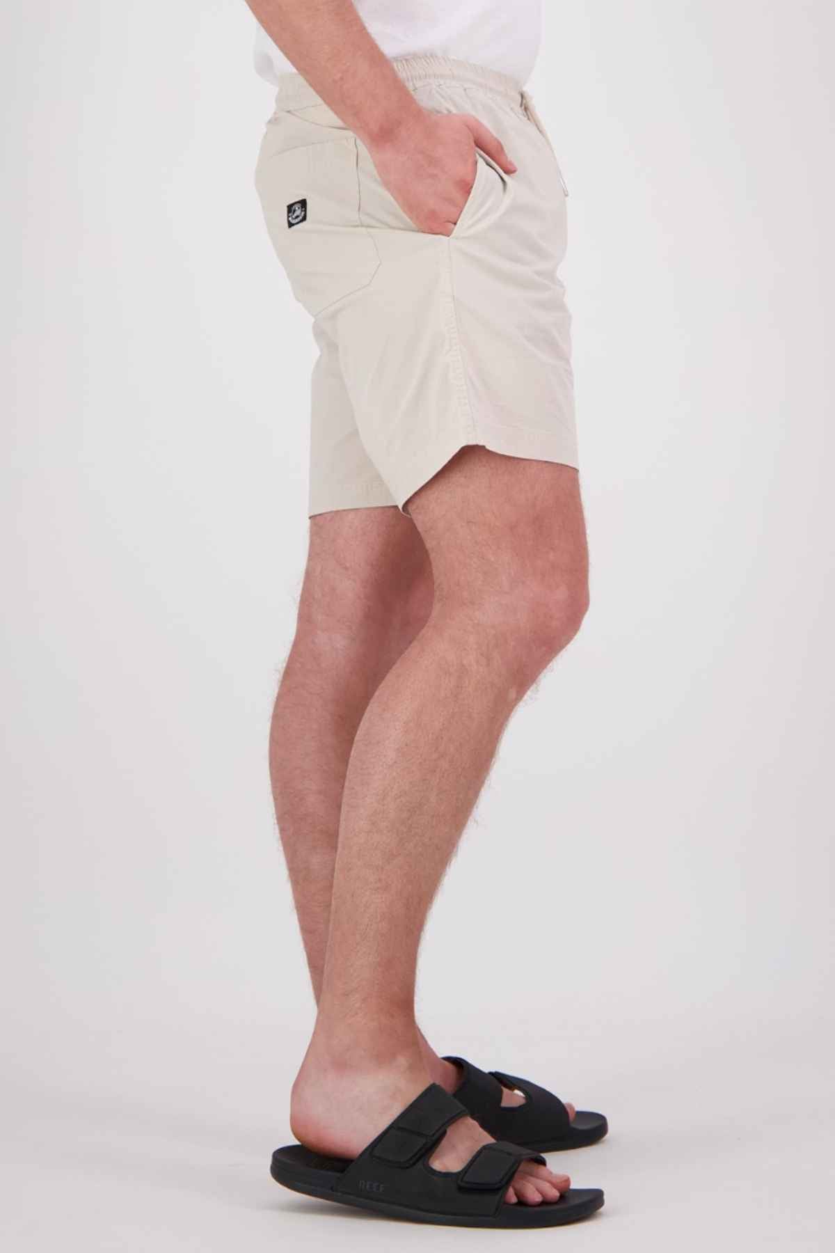 A person dressed in beige Swanndri Frankton Shorts, made from a comfortable cotton blend, along with black slide sandals stands sideways with their hands in the pockets against a simple background. The shorts have an elastic waist and feature a small logo on the pocket.