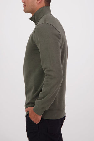 Wearing the eco-friendly Swanndri Fundamentals Quarter Zip in olive green and black pants, a person stands in profile with hands in pockets against a plain white background.