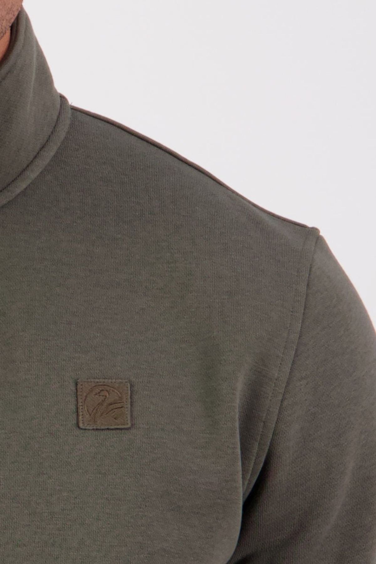 Close-up of a person wearing the Swanndri Fundamentals Quarter Zip in olive green, crafted from recycled polyester with a small rectangular bird logo patch on the chest. The background is plain and light-colored.