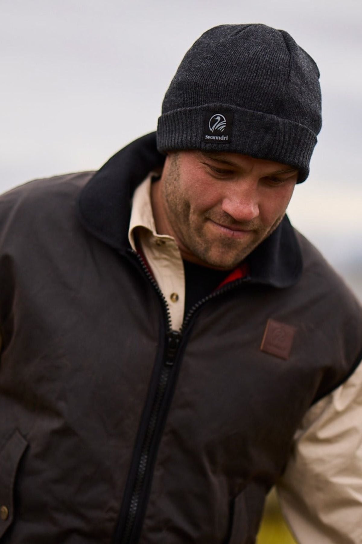A man wearing a Swanndri Grafton Beanie and a brown jacket over a beige shirt looks down with a slight smile outdoors on a cloudy day.