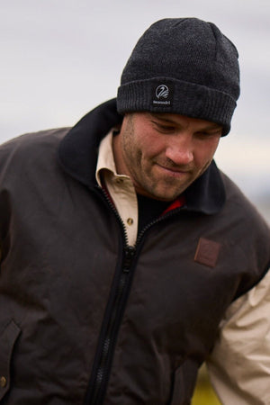 A man wearing a Swanndri Grafton Beanie and a brown jacket over a beige shirt looks down with a slight smile outdoors on a cloudy day.