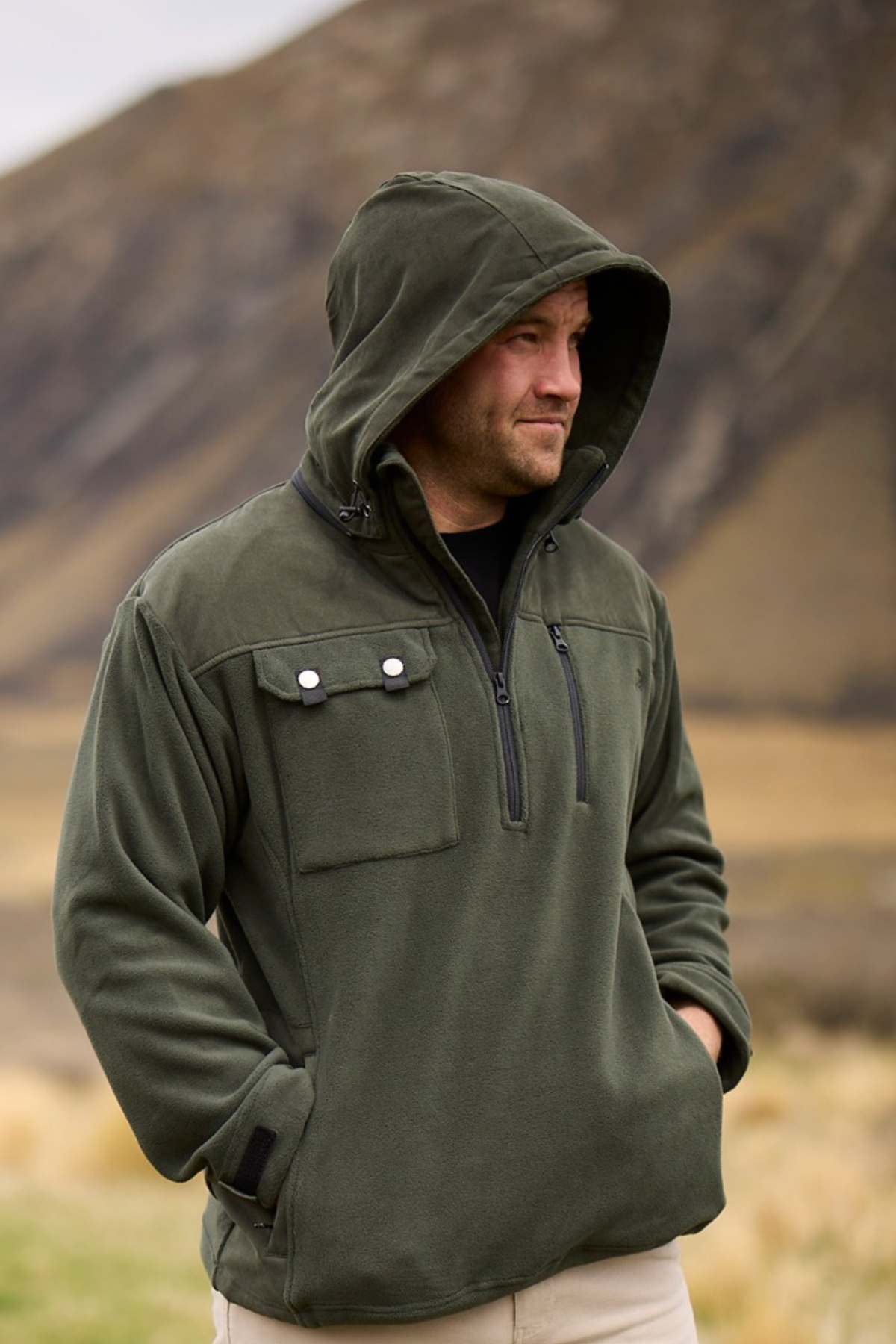 A man dressed in a Swanndri High Rock Fleece Pullover stands outdoors in a mountainous area. He has his hands in the pockets of his olive green hooded jacket, and he is looking off to the side. The background features blurred, rocky hills and dry grass.
